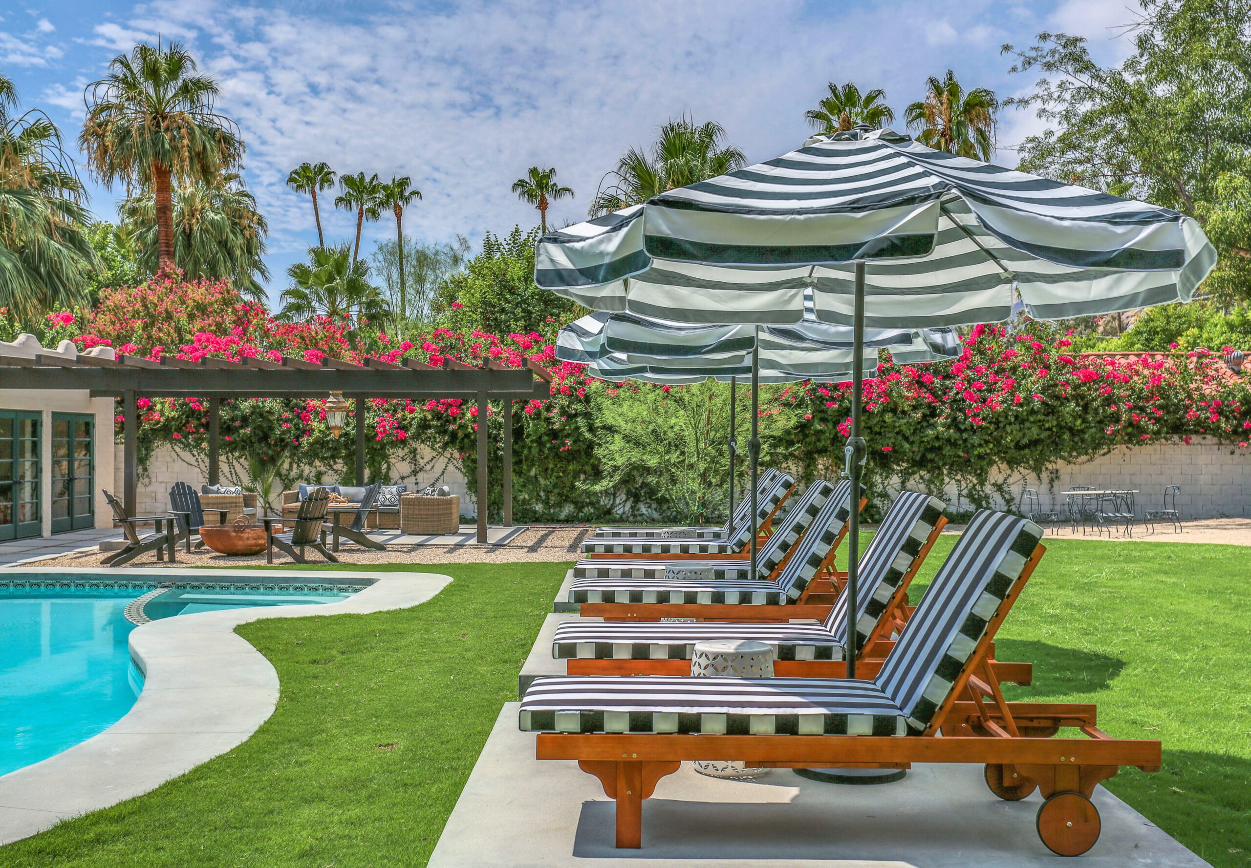 Four striped lounge chairs and umbrellas next to a pool, surrounded by green grass, trees, and pink flowers under a partly cloudy sky.
