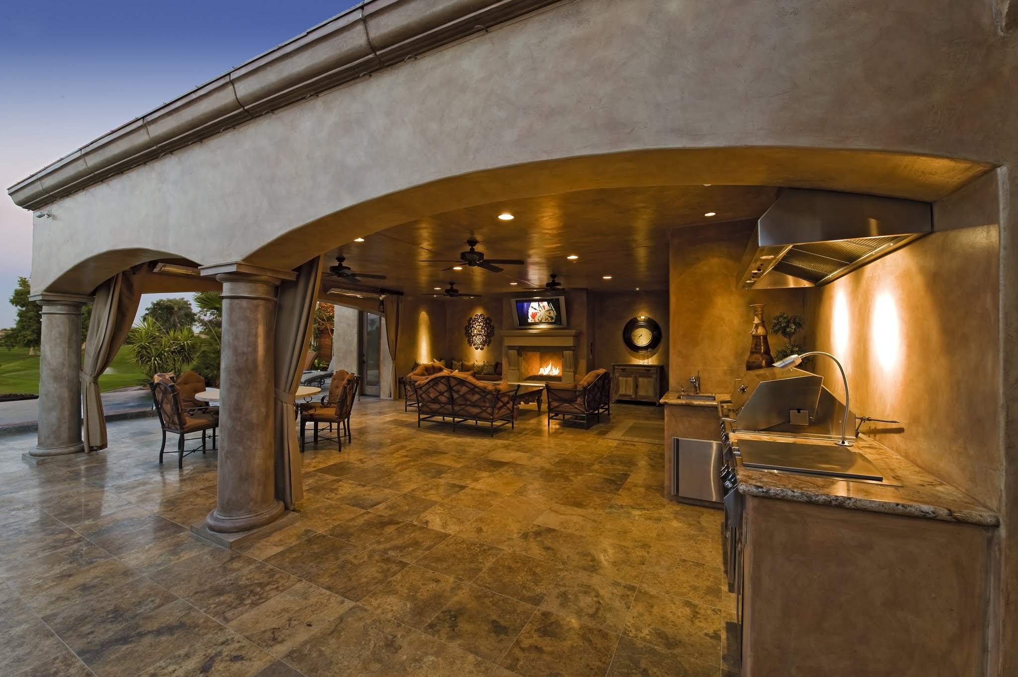 Outdoor kitchen and living area with stone flooring, ceiling fans, and elegant columns. Includes a TV, grill, and seating, with a view of a garden through the arches.
