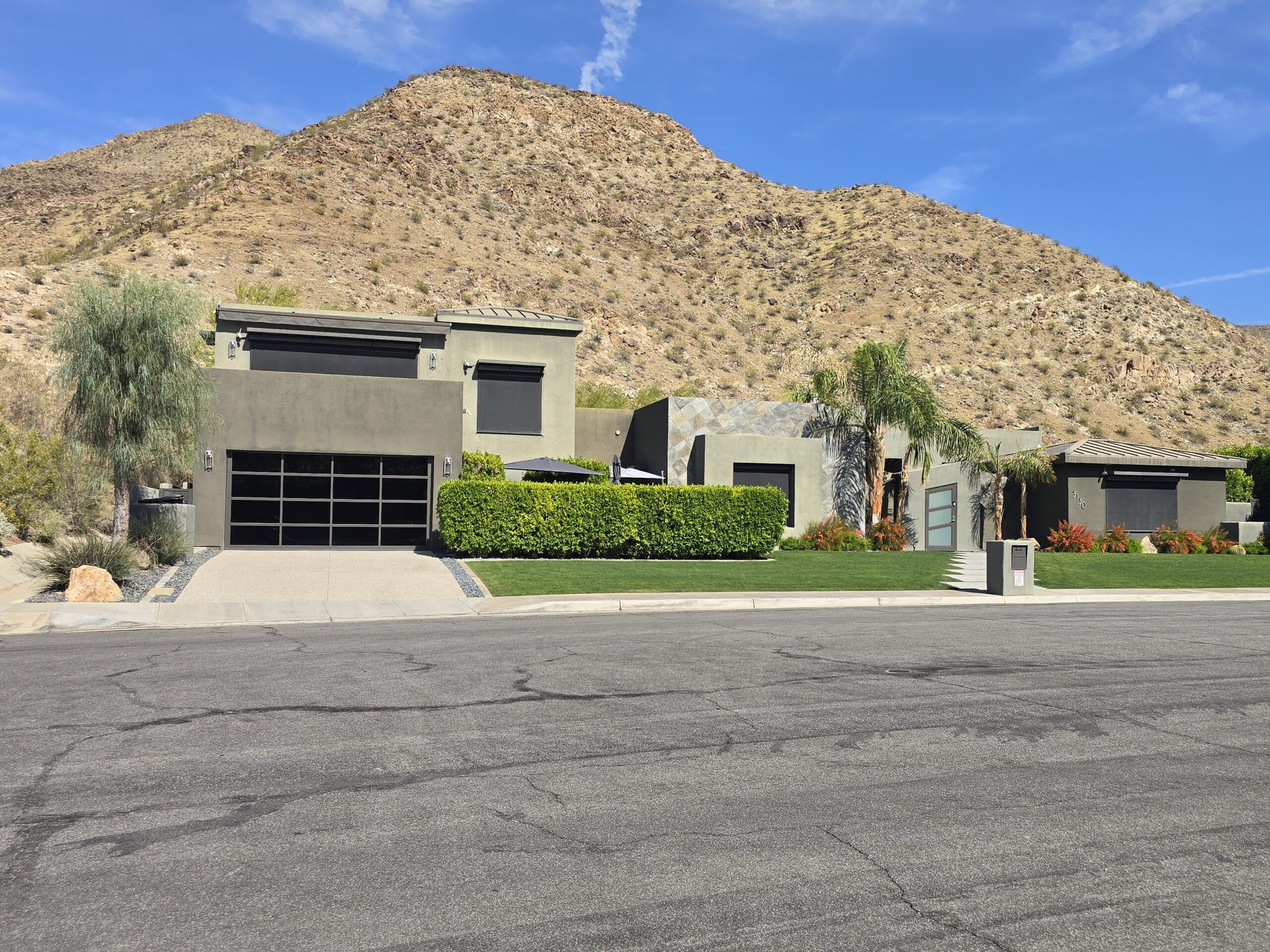 Modern gray house with a flat roof, garage, and well-maintained lawn, set against a backdrop of arid, rocky hills under a clear blue sky.