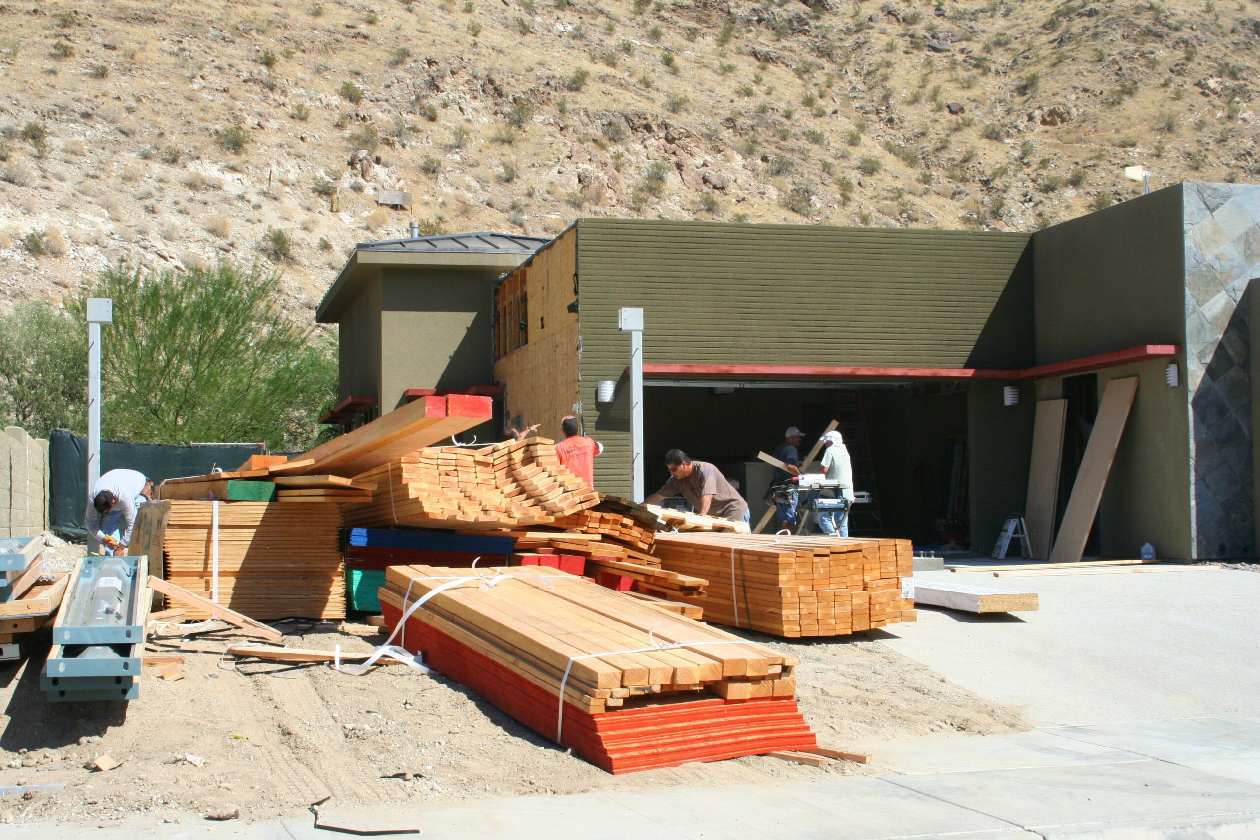 Construction site with stacks of wood and materials in the foreground, and workers building a modern house against a rocky hillside backdrop.