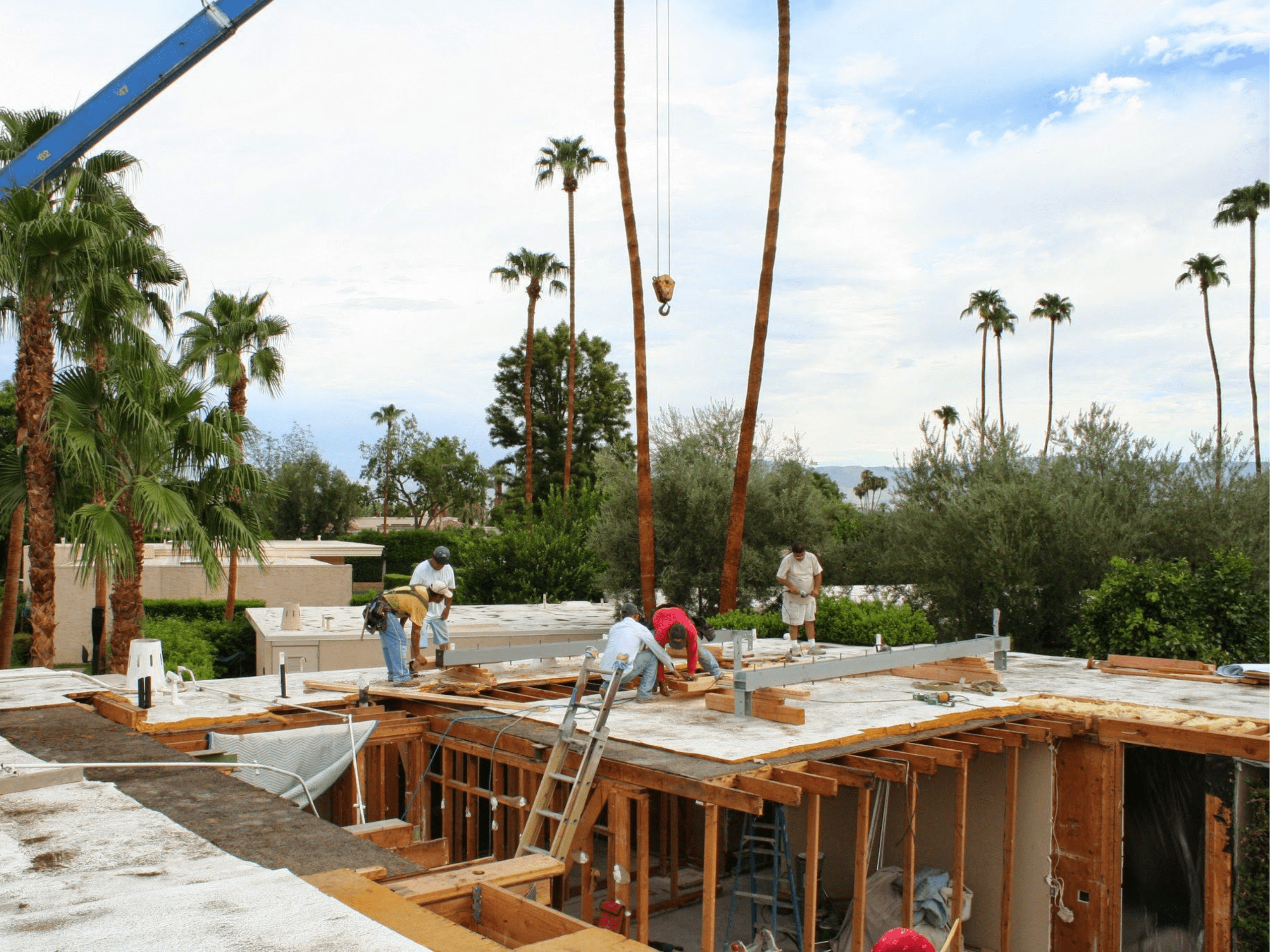 Construction workers building a house with palm trees in the background. A crane is visible, and several people are working on the wooden framework.