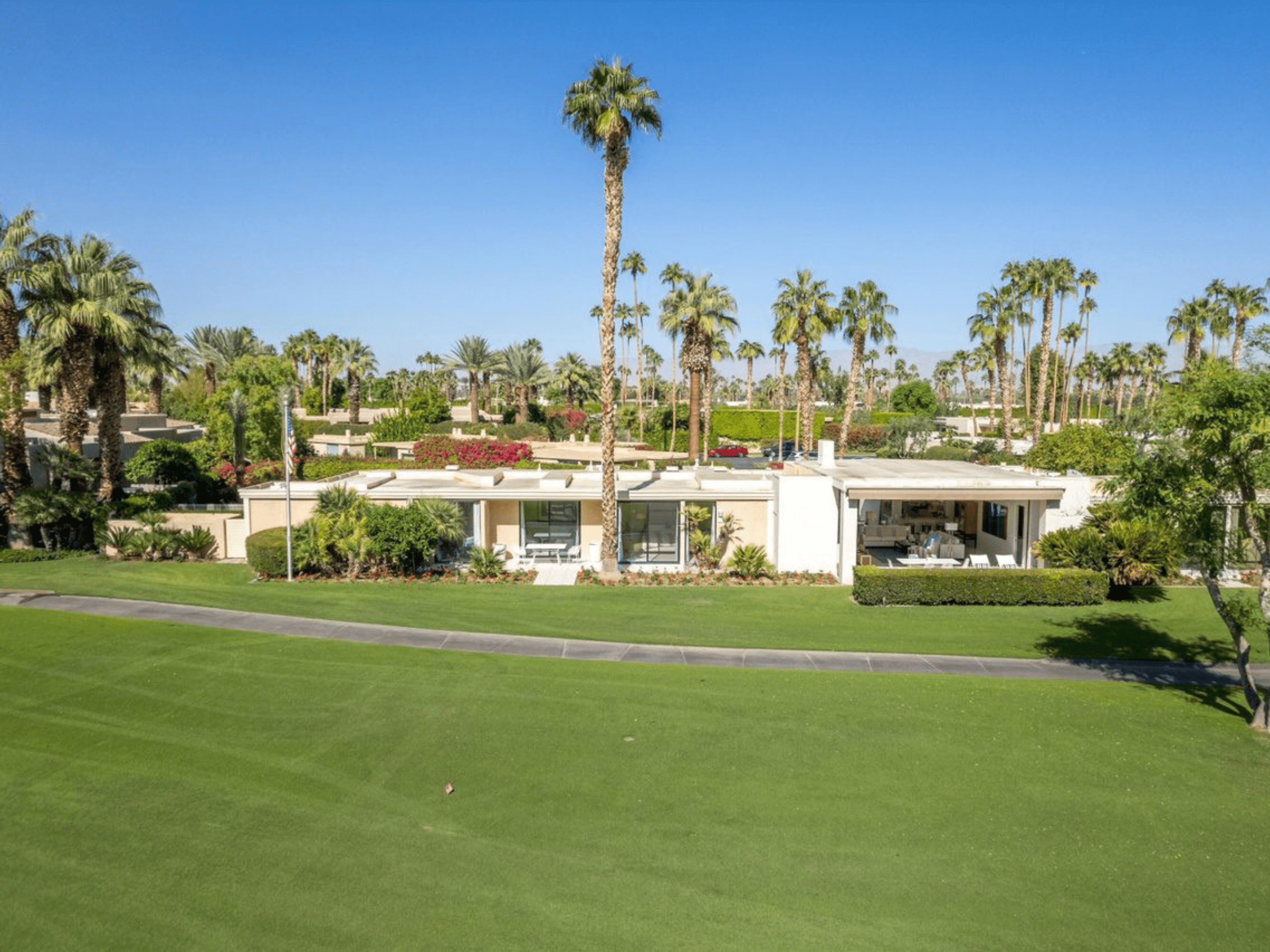A single-story house with large windows is surrounded by palm trees and greenery, adjacent to a well-maintained lawn under a clear blue sky.