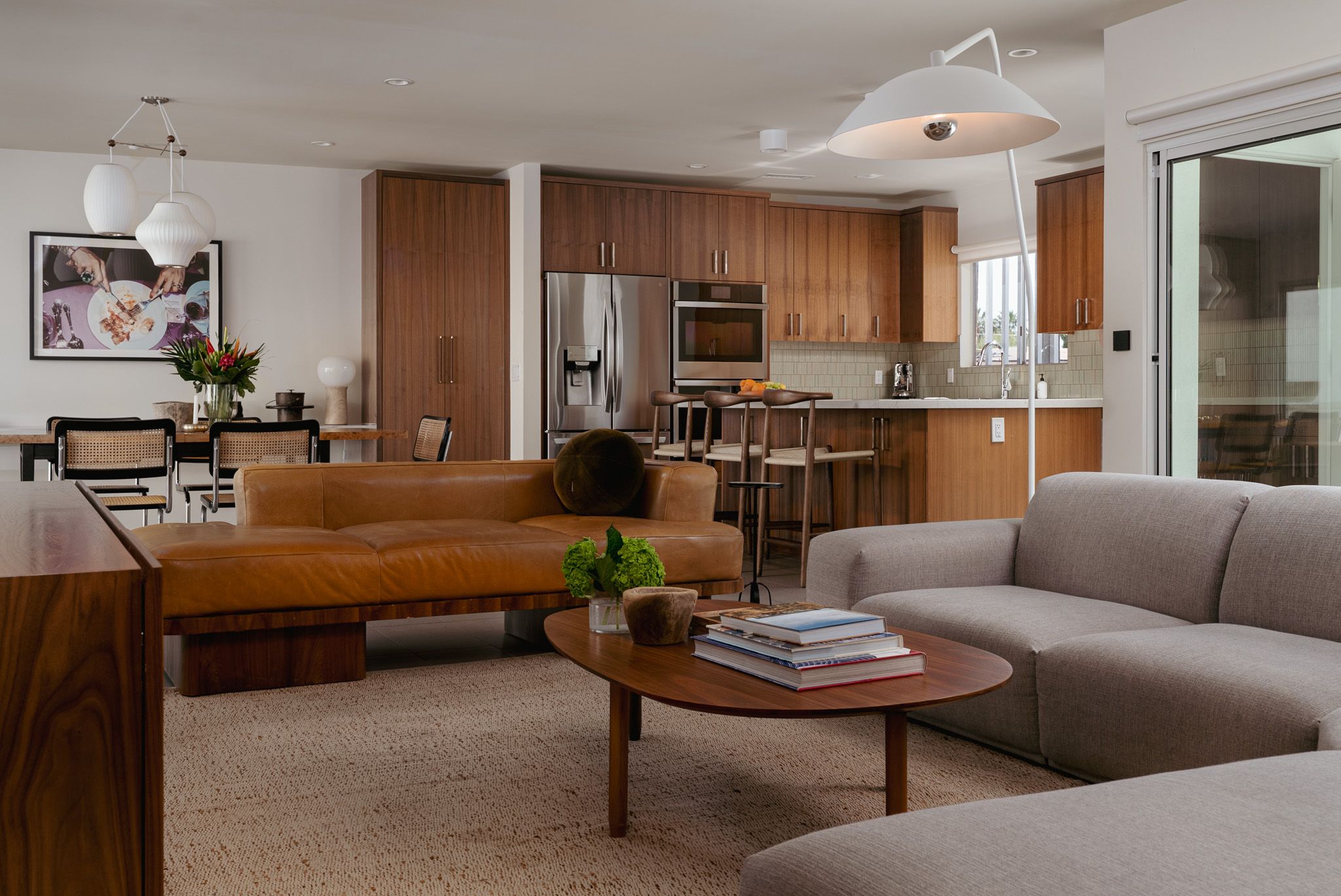 Modern living room with a brown leather sofa, gray sectional sofa, wooden coffee table, and kitchen in the background. Large windows and a contemporary light fixture enhance the space.