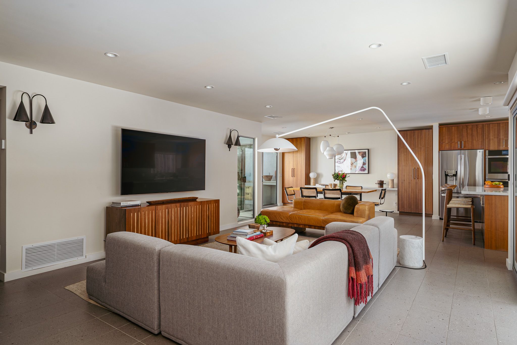 Modern living room featuring a large gray sofa, flat-screen TV, wooden cabinet, arched floor lamp, and view into an open kitchen and dining area with a brown leather bench.