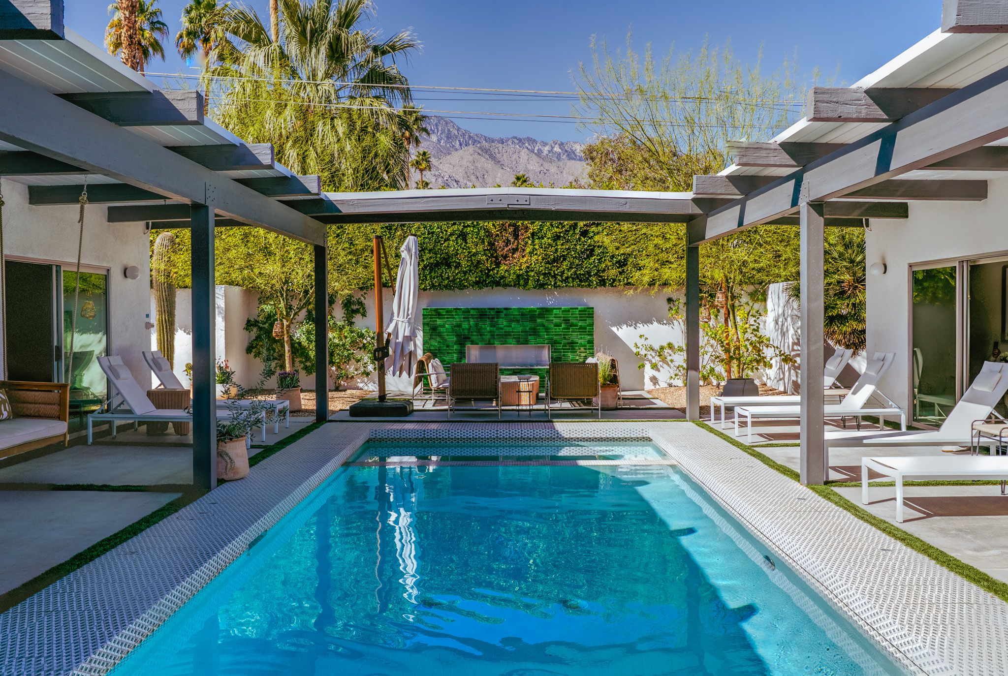 Modern outdoor pool area with lounge chairs, patio furniture, and shaded pergola. Surrounded by lush greenery and palm trees under a clear blue sky.