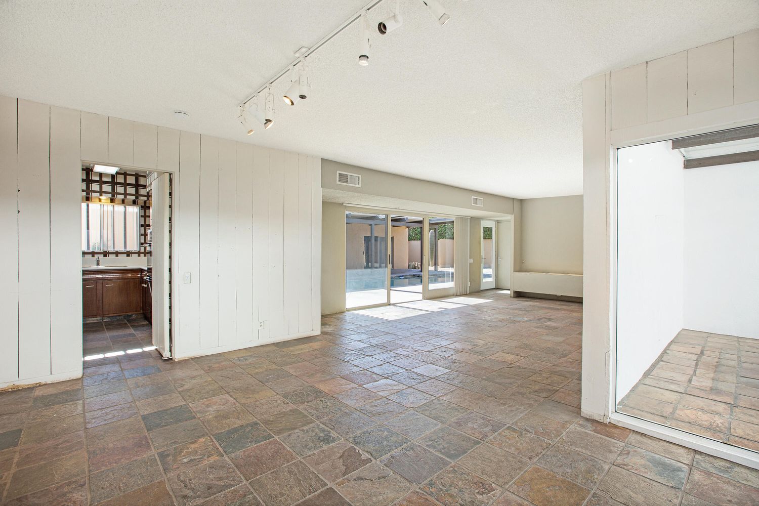 Spacious room with tiled floor, white walls, track lighting, and large sliding glass doors leading to an outdoor area. An open doorway connects to a bathroom on the left.