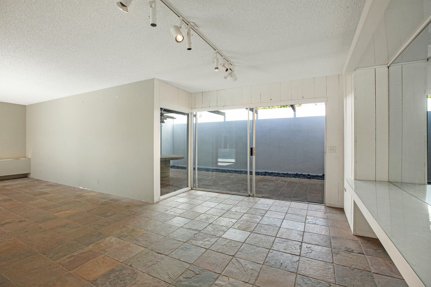 Empty room with tiled floor, track lighting, and sliding glass doors leading to an outdoor area. White walls and ceiling, with a small built-in mirror on the right.