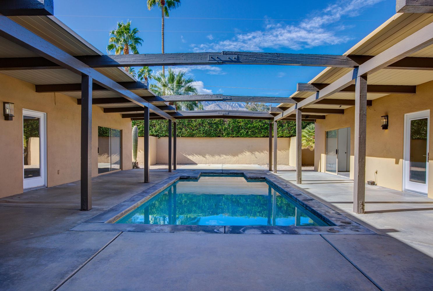Rectangular swimming pool under a partially covered patio with wooden beams, surrounded by beige walls and glass doors. Palm trees and a blue sky are visible in the background.